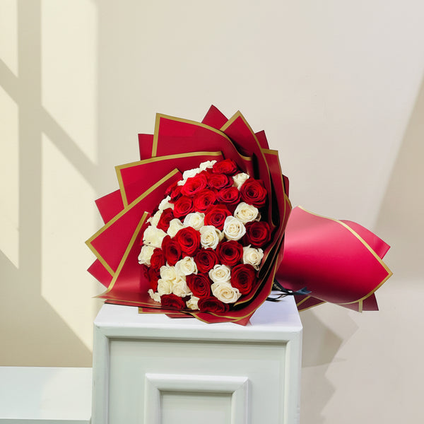 A large bouquet of red and white roses wrapped in red paper with gold edges, displayed on a white pedestal against a plain light background.