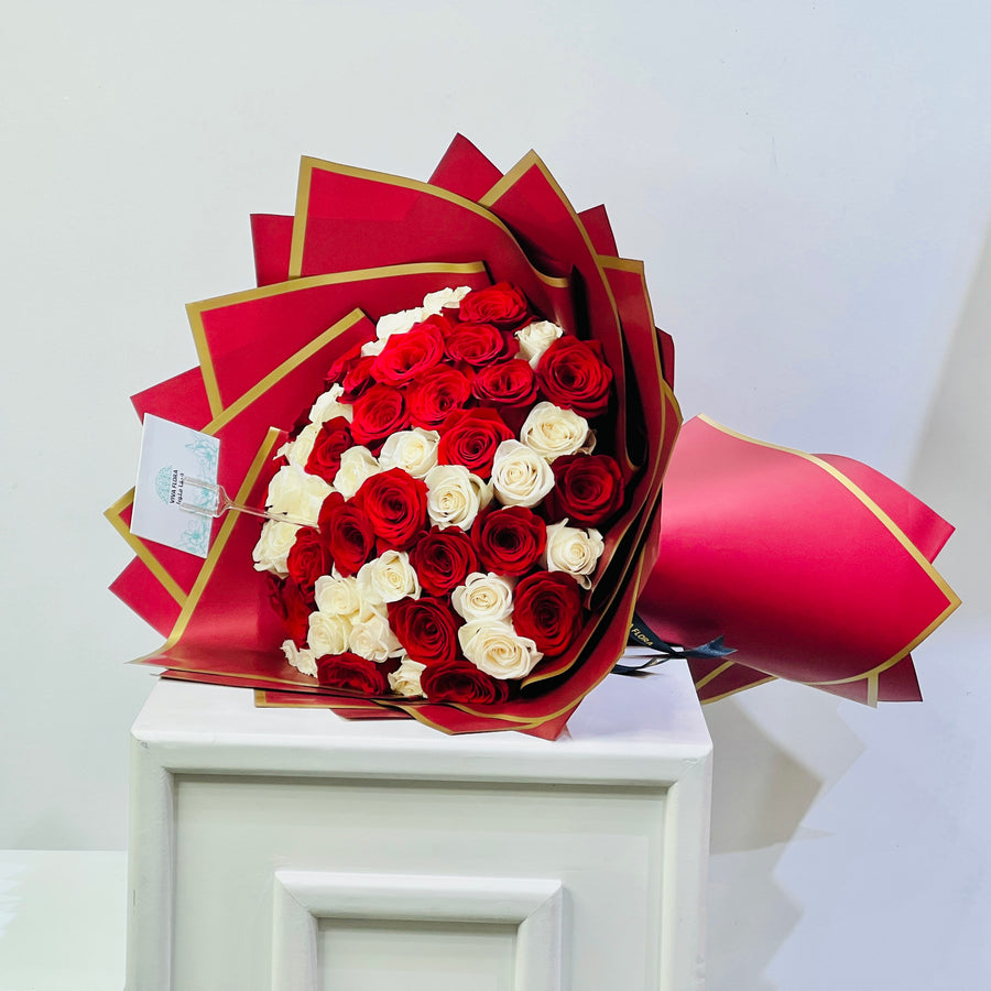 A large bouquet of red and white roses wrapped in red paper with gold edges, displayed on a white pedestal against a plain light background.