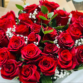 A large bouquet of deep red roses with green leaves and small white baby's breath, wrapped in white tissue paper and clear cellophane, held in front of a shop window.