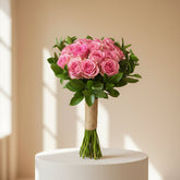 A person wearing a black abaya and headscarf holds a large bouquet of pink roses with green leaves, wrapped in twine at the stem, against a softly blurred indoor background.