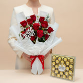 A bouquet of 15 red roses wrapped in white tissue paper with a red ribbon and a 'With Love' sign, displayed next to a box of Ferrero Rocher chocolates on a round pedestal against a warm beige background.