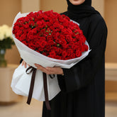 A large bouquet of red roses wrapped in white paper with a black ribbon bow, displayed on a round white table against a warm beige background.