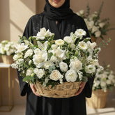 A large white floral arrangement in a woven basket, featuring white roses, carnations, daisies, and green foliage, displayed on a white pedestal against a beige wall with soft window shadows.