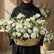 A large white floral arrangement in a woven basket, featuring white roses, carnations, daisies, and green foliage, displayed on a white pedestal against a beige wall with soft window shadows.
