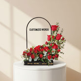 A floral arrangement of red roses and green foliage in a black rectangular planter on a white pedestal, with a black arched sign behind it reading 'CUSTOMIZED WORDS'.