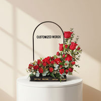 A floral arrangement of red roses and green foliage in a black rectangular planter on a white pedestal, with a black arched sign behind it reading 'CUSTOMIZED WORDS'.