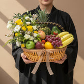 A woven wicker basket filled with assorted fruits and flowers, displayed on a round white pedestal against a warm beige background. The basket contains yellow bananas, red grapes, green apples, a pineapple, and white and yellow flowers tied with a sheer ribbon.