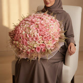 A large, dense floral bouquet made up of mostly pink roses with cream and blush tones, wrapped in pale pink paper and held by a qatar lady who is sitting in on a white chair 