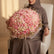 A large, dense floral bouquet made up of mostly pink roses with cream and blush tones, wrapped in pale pink paper and held by a qatar lady who is sitting in on a white chair 