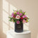 A vibrant floral arrangement of pink, red, and light pink roses with green foliage, presented in a black cylindrical box with gold branding, set against a plain white background.