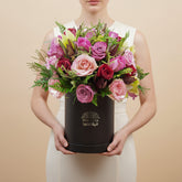 A vibrant floral arrangement of pink, red, and light pink roses with green foliage, presented in a black cylindrical box with gold branding, set against a plain white background.
