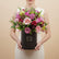 A vibrant floral arrangement of pink, red, and light pink roses with green foliage, presented in a black cylindrical box with gold branding, set against a plain white background.