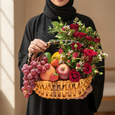 A wicker basket filled with red roses, grapes, strawberries, and fruit, displayed on a white cylindrical pedestal against a warm beige background with soft window-shadow lighting.