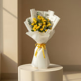 A person wearing a black abaya and black hijab holds a large bouquet of yellow roses wrapped in white paper with a yellow ribbon, against a warm beige background.