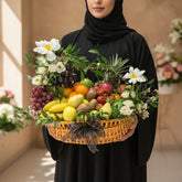 A woven wicker basket filled with assorted fruits and white flowers, displayed on a white cylindrical pedestal against a warm beige background with soft window-shadow lighting.
