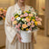 A vibrant floral arrangement featuring pink, peach, yellow, and white flowers with green foliage, presented in a white round box with the brand name 'VINE FLORIST' printed on the front, set on a white pedestal against a warm beige background.