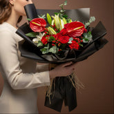 A vibrant flower bouquet featuring red anthuriums, red roses, green lily of the valley, and green eucalyptus leaves, wrapped in black paper and tied with a natural jute bow, displayed on a white pedestal against a warm beige background.