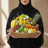 A woven basket filled with assorted fresh fruit and flowers, displayed on a white round pedestal against a warm beige background with soft window-shadow lighting.