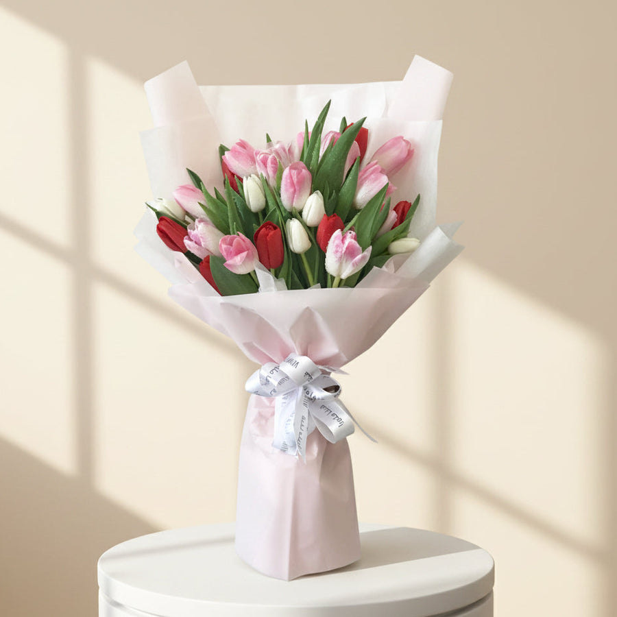  a wrapped bouquet of pink, red, and white tulips with green leaves and stems, tied with a white ribbon, in a softly lit indoor setting with blurred shelves in the background.