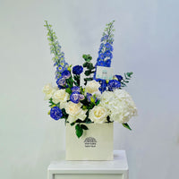 A floral arrangement in a cream-colored square box, featuring blue delphiniums, white and blue roses, and white hydrangeas, displayed on a white pedestal against a light gray background.