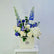 A floral arrangement in a cream-colored square box, featuring blue delphiniums, white and blue roses, and white hydrangeas, displayed on a white pedestal against a light gray background.
