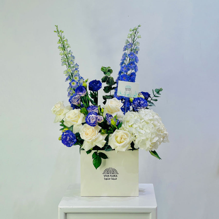 A floral arrangement in a cream-colored square box, featuring blue delphiniums, white and blue roses, and white hydrangeas, displayed on a white pedestal against a light gray background.
