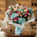 A vibrant bouquet of artificial flowers wrapped in white tissue paper and tied with a light blue ribbon, placed on a gray tiled floor in front of a blurred shop background.