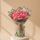 A person in a black abaya holds a large bouquet of pink roses wrapped in sheer gray paper with black trim and a tan raffia tie, against a softly blurred beige indoor background with flowers on shelves.