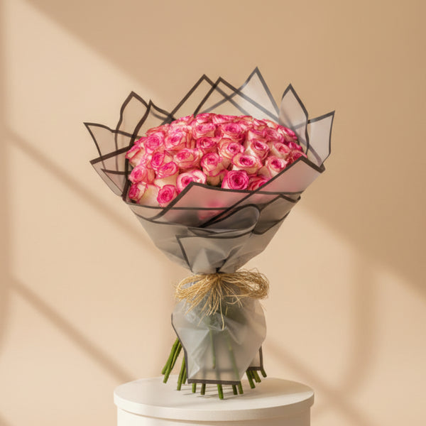 A person in a black abaya holds a large bouquet of pink roses wrapped in sheer gray paper with black trim and a tan raffia tie, against a softly blurred beige indoor background with flowers on shelves.