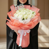 A bouquet of white roses wrapped in sheer coral and white paper with a white ribbon, displayed on a white pedestal against a warm beige background with soft window shadows.