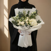 A bouquet of white flowers and green foliage wrapped in white tissue paper and tied with a white ribbon, displayed on a white pedestal against a warm beige background with soft window shadows.
