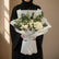 A bouquet of white flowers and green foliage wrapped in white tissue paper and tied with a white ribbon, displayed on a white pedestal against a warm beige background with soft window shadows.