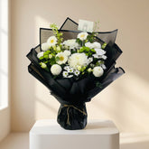 A bouquet of white flowers, including daisies and carnations, wrapped in black paper and tied with a thin brown string, displayed on a white pedestal against a white wall.