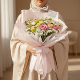 A small floral bouquet wrapped in pale pink tissue paper and tied with a matching ribbon, displayed on a round white pedestal against a warm beige background with soft window-shadow lighting.