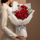 A bouquet of red roses wrapped in white tissue paper with a silver ribbon and a white script card reading 'Love', displayed on a white pedestal against a warm beige background with soft window shadows.