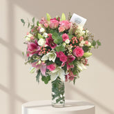 A vibrant floral arrangement of pink roses, lilies, and white orchids in a clear glass vase, displayed on a round wooden-topped table in a modern living room setting.