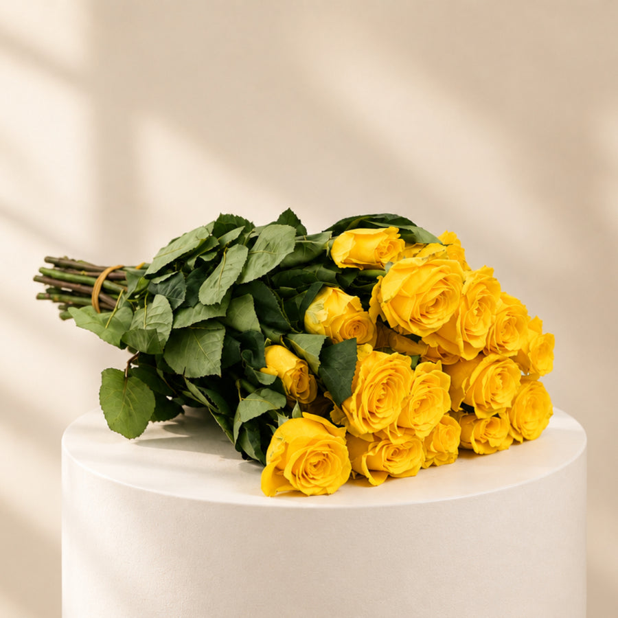 A bouquet of yellow roses with green leaves and stems, tied together with a rubber band, resting on a plain white surface.