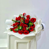 A bouquet of red roses with green leaves, wrapped in white tissue paper and a large white bow, displayed on a white pedestal against a plain light background.