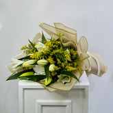 A floral bouquet with white lilies, yellow-green filler flowers, and green leaves, wrapped in beige paper and tied with a matching ribbon, displayed on a white cabinet against a light gray wall.