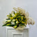 A floral bouquet with white lilies, yellow-green filler flowers, and green leaves, wrapped in beige paper and tied with a matching ribbon, displayed on a white cabinet against a light gray wall.