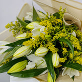 A close-up of a fresh flower bouquet featuring white lilies, yellow-green buds, and dense clusters of small yellow flowers, wrapped in light tan paper.
