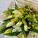 A close-up of a fresh flower bouquet featuring white lilies, yellow-green buds, and dense clusters of small yellow flowers, wrapped in light tan paper.