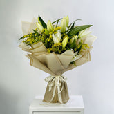 A fresh flower bouquet featuring white lilies, yellow-green filler flowers, and green leaves, wrapped in beige paper and tied with a cream ribbon, displayed on a white pedestal against a light gray background.