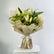 A fresh flower bouquet featuring white lilies, yellow-green filler flowers, and green leaves, wrapped in beige paper and tied with a cream ribbon, displayed on a white pedestal against a light gray background.