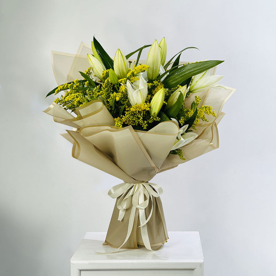A fresh flower bouquet featuring white lilies, yellow-green filler flowers, and green leaves, wrapped in beige paper and tied with a cream ribbon, displayed on a white pedestal against a light gray background.
