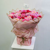 A large bouquet of pink, red, and peach flowers wrapped in light pink paper with gold geometric lines, tied with twine and standing on a white surface against a plain light gray wall.