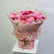 A large bouquet of pink, red, and peach flowers wrapped in light pink paper with gold geometric lines, tied with twine and standing on a white surface against a plain light gray wall.