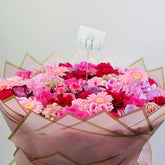 A large, vibrant bouquet of assorted pink, red, and purple flowers wrapped in pink paper with gold accents, featuring a small decorative card on a white background.