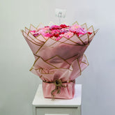 A bouquet of pink carnations wrapped in pink paper with gold geometric patterns, tied with twine and displayed on a white pedestal against a light gray wall.