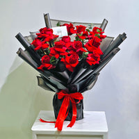 A large bouquet of red roses wrapped in black paper and tied with a red ribbon, displayed on a white table against a plain light gray wall.